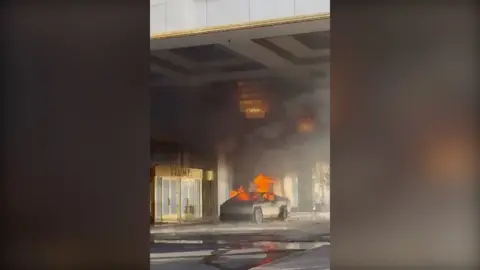 A Tesla truck has large flames emerging from it outside a golden doorway with "TRUMP" emblazoned above it.