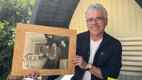 A man with gray hair wearing glasses smiles as he holds an old black and white photograph on a cardboard mount with signatures around it. The photo shows him as a child with Graham Taylor, a dark-haired man wearing a blazer and tie, and a taller man with a dark mullet wearing a tie and jumper.