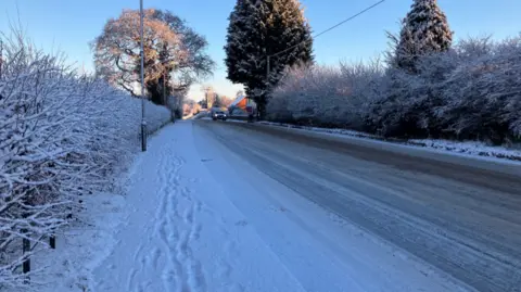 A road and pavement covered in a layer in snow. There is a vehicle approaching with its lights on