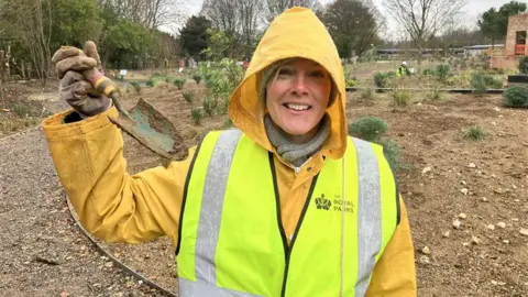 Sonya, wearing a grey hat and scarf underneath a yellow raincoat and Royal Parks hi vis waistcoat, holds up a muddied trowel and smiles