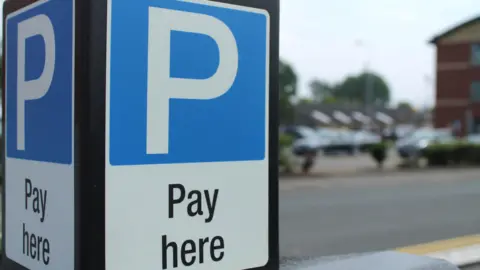 A blue and white sign with a blue parking symbol on it. In the background appears to be a road and parked cars beside a building.