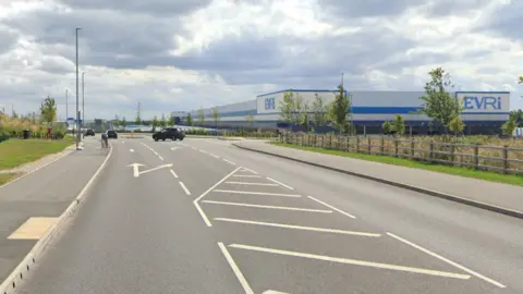 Google Street View image of a new-looking road with an industrial estate on the right. There is a large Evri dispatch factory. A car is pulling out of the road on the right.