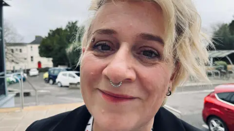 The image shows an outdoor setting with a paved area in the foreground and a car park in the background. The person in the foreground is wearing a black jacket and a white lanyard. Their hair is light-coloured and styled in loose waves. Several cars are parked, including a white car, a yellow car, and a red car. Behind the car park, there are trees and a light-coloured building with multiple windows and a bright red door.