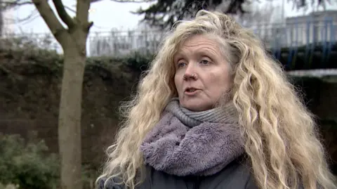 Cathy Scott dressed in a grey coat wearing a purple fluffy scarf stood in a park