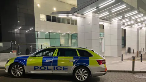 A police car parked outside the Royal Victoria Hospital Emergency Department. The building is white. The police car is yellow and blue and says POLICE along the side of it.