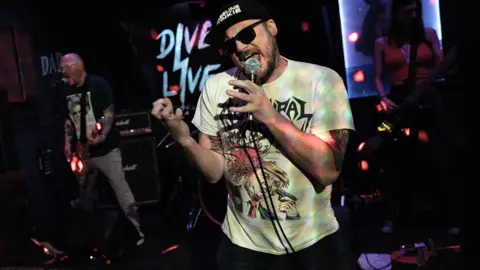 The band Meth Club 7 appear on stage at the Dive in Wolverhampton. The lead singer wears a black cap, dark glasses and is singing into a microphone. Behind him other band members, one female and one male, can be seen on state