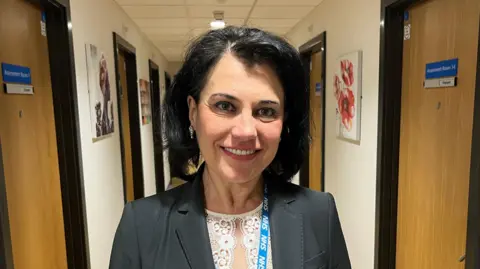 Consultant Psychiatrist Vesna Jordanova stands in the corridor at Albion Place, hub for the Kent & Medway Mental Health Trust. She has shoulder length black hair and is wearing a grey suit jacket and a lacey white top. She has a blue lanyard on