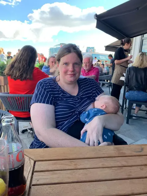 Louise Owen Louise is sat at an outdoor cafe breastfeeding. There are other customers in the background.