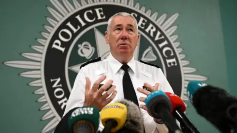 Getty Images Shows Jon Boutcher in a white shirt and black tie standing in front of the Police Service of Northern Ireland crest. 