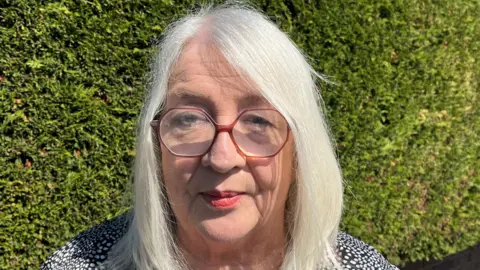 Gaynor Hughes photographed standing outside in front of a green hedge. She has silver mid length hair, has red lipstick and wears glasses. 