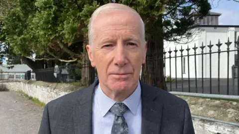 A man stands close to the camera, with short white hair, wearing a grey suit and blue shirt.