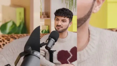 Shabaz Ali has dark hair and is wearing a beige jumper