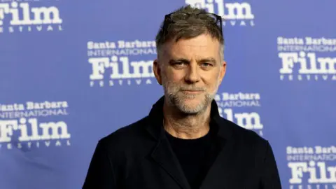 Getty Images A white haired man with glasses on his head and a white beard, standing in front of a blue backdrop saying Santa Barbara Film Festival. 