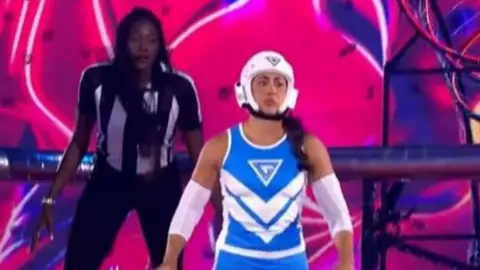 Keeva Nelly in a blue and white leotard and skullcup standing on a pink stage with pink and purple background