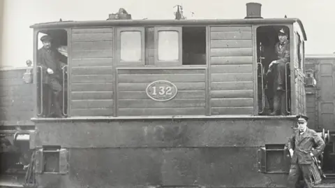 The Board of Trustees, Science Museum Group A black and white photograph of a tram engine which is in the middle. Two people are standing in the doorways of the engine either side and a person is stood on the ground on the right. 