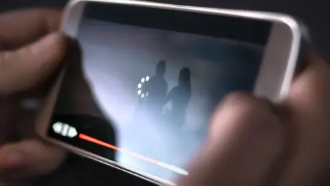 Getty Images A close-up of someone's fingers holding up a phone, while watching a film. It has a circle of dots in the middle of the picture, indicating it is buffering. 