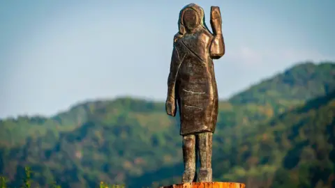 A bronze replica depicting US First Lady Melania Trump waving her left hand pictured in a field near the US First Lady's hometown Sevnica. The statue looks rustic in style, with no visible facial features.