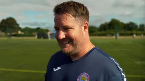 Jonny Michel/BBC Kevin Fryer smiles while looking to the left of the camera. He is wearing a football shirt and is standing on a football pitch. He has short brown hair.