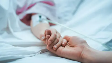 Getty Images A child in a hospital bed holding an adult's hand. 