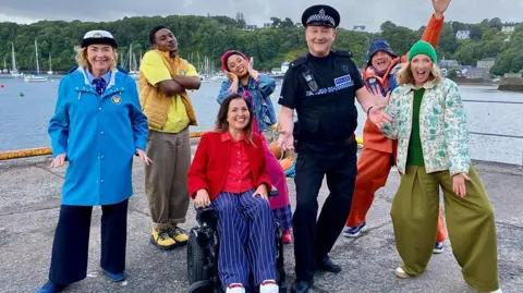 Lion TV/MediaScotland BBC Seven people are pictured on a pier with the sea behind. There are lots of yachts in the water. Six of the characters are wearing different colours and have their hands out, one is seated, they are all smiling.