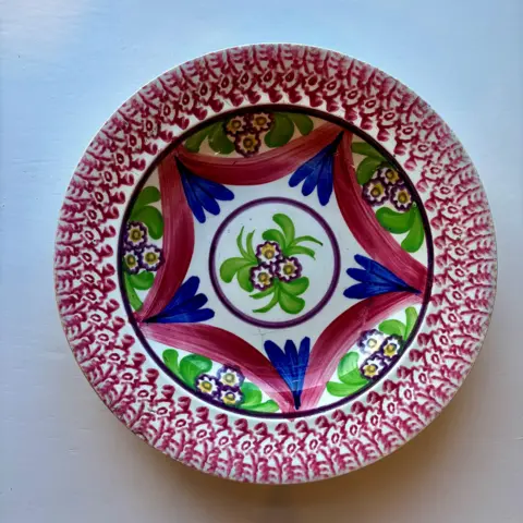 Scottish Maritime Museum A brightly coloured decorative bowl, with red flowers around the rim of the plate, and a green, red, blue and yellow floral pattern on the inside. Three daisies are painted within a circle in the centre.