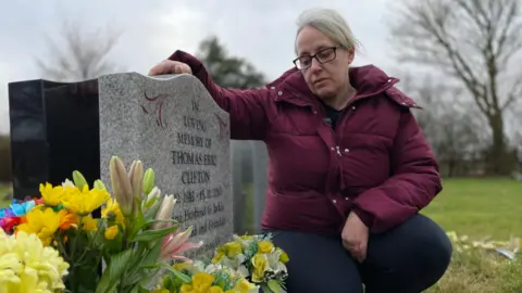 Jo Makel/BBC A woman kneels down next to the headstone of a grave. She rests her right arm on the headstone. It is the grave of her father. She is looking at the headstone. She has blonde hair and dark-rimmed glasses. She is wearing a burgundy coat and black trousers.
