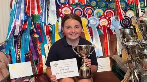 A 10-year-old girl with brown hair and brown eyes is smiling at the camera. She is sitting on her sofa and holding up a certificate and a trophy. Her hair is tied in a ponytail and she is wearing a black polo shirt. Draped all over the sofa are brightly coloured rosettes - more than 50 of them. To the right side of the photo there are 20 polished silver trophies.