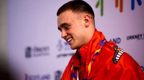 EUAN DUFF Harry Robinson, who has short brown hair, smiling. He is wearing a red top and has a red Manx flag draped over his shoulders.