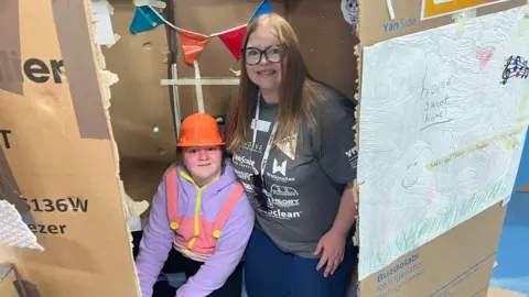 A nine-year-old girl wearing an orange hard hat and a colourful purple hoodie with a dungaree pattern on smiles as she sits beside her mum in a large cardboard box. The cardboard box has been decorated for them to spend the night in. Her mum is also smiling, has long, straight, dirty blonde hair and glasses, and is wearing blue leggings and a grey T-shirt with white company logos on it.