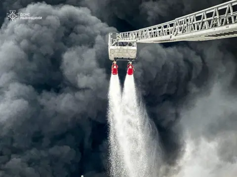Thick dark grey smoke billows from the Brody oil hub as firefighters tackle an unseen fire with two hoses from an extended ladder.
