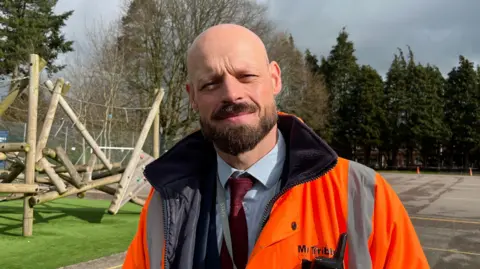 A man standing in a school playground wearing a bright orange jacket