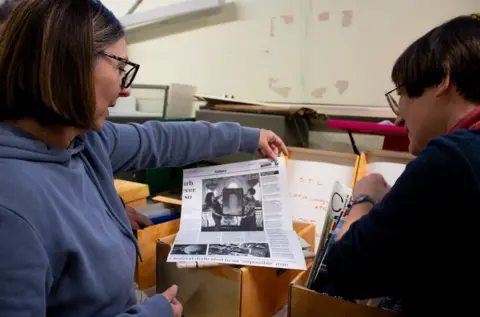 Puffin Alwarey Two women are looking through boxes of archive material. One is holding up a newspaper cutting. They both have short hair and are wearing glasses. 