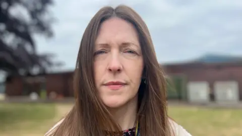Fran Rhodes has long brown hair and is wearing a white fluffy coat. The school field behind her has been blurred.