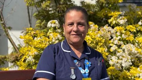 Susan Lesbirel. She has dark tied back hair and is wearing a blue nurses uniform.