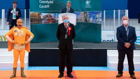 Getty Images Then First Minister of Wales Mark Drakeford (centre) looks on after winning the Cardiff West constituency as candidate Captain Beany (L) looks on at the Cardiff House of Sport on May 7, 2021