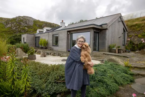 IWC Media Susie stands in a landscaped garden holding a small dog, in front of a modern, wood-clad house set against rolling hills.
The home features large windows, clean lines, and a mix of rustic and contemporary design elements.
Surrounding plants, stone paths, and a cloudy sky create a calm, natural countryside setting.