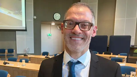 Adam Zerny with short grey hair and beard, wearing brown-framed glasses, a white shirt, grey tie and black jacket. He is standing in front of long wooden tables in a council chamber.