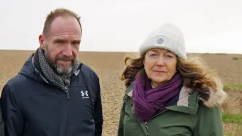 Ralph Fianna and fellow campaigner Fiona Gilmore painted the beach in Eldeburg in Safok. They have short, light brown hair and a brown beard, wearing a black pointed jacket and black and white dupatta. Fiona has long, wavy, obnour hair, a white woolen cap, purple scarf and khaki wearing green waterproof coat, with a fur-line hood. The sky is cloud and they are standing on the shingle.