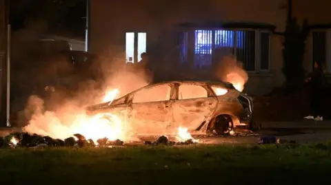 Getty Images A car set aflame in front of a lit up house, a grass verge can be seen before the car. A silhouette of a person spraying water on the car.