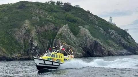 St John Ambulance and Rescue Service Guernsey Flying Christine III at sea off Sark. It is making a large wash. The land behind it is steep and rocky and there is a lighthouse just visible.
