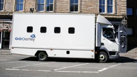 A GEOAmey van driving through the Pleasance area of Edinburgh. The van is white with four dark windows mid way up the vehicle. The name of the company and a Scotland flag, a white cross on a blue background, is visible on the vehicle. It is driving in front of a light-stone tenement building.