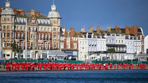 Line of runners in red Santa outfits making their way along Weymouth Beach with the sea to the right and buildings along the left.