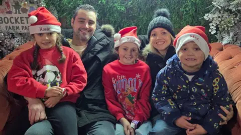 Supplied Parents Karen and Adam sit with their three children on a sofa. The three children are wearing Christmas hats and everyone is smiling for the camera.