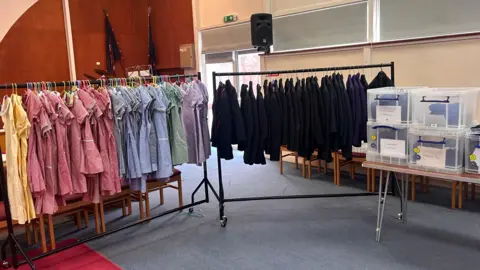 In a hall, there are two rails of clothes. On one are summer gingham dresses in different colours, and on the other, black blazers. Next to them on a long table are clear boxes.