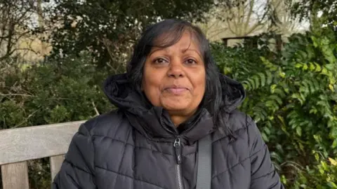 Louise Parker/BBC Helen Newton is sitting on a bench with hedges in the background. She is wearing a black puffer jacket. 