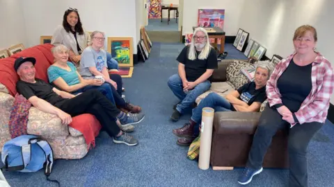 Seven people sitting on two sofas in a white-walled room with paintings on the floor leaning on the walls.
(Left to right) Barry Greenwood, Caro Blount-Shah, Ann Driver, Lauren Kelly, Ken Woods, Cath Muldowney, Pauline Cooke