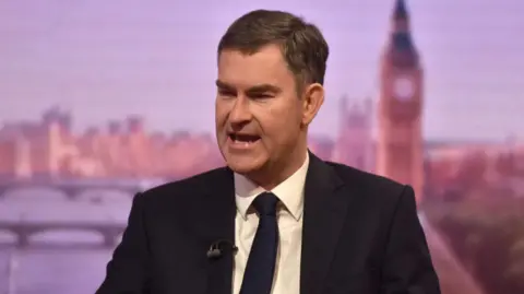 David Gauke wearing a black suit, a black tie and a white shirt. He sits in a BBC studio and is talking to the interviewer.