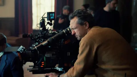 A24 Lol Crawley, who has short greying hair and is wearing a brown jumper, looking through the lens of a large black camera which is pointed at something off the left side of the shot. In the background is a window with red curtains, and various film crew members dressed in black.