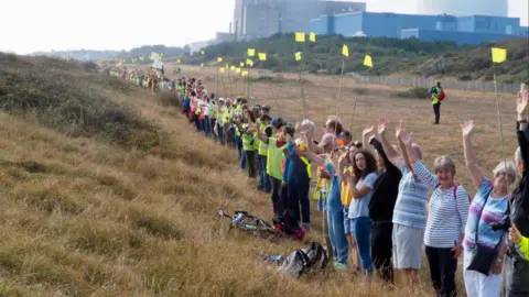 Stop Sizewell C/Brian Lowry Hundreds of people line a coastal path in front of the Sizewell nuclear power stations. Some of them hold small yellow flags as they wave at the camera.