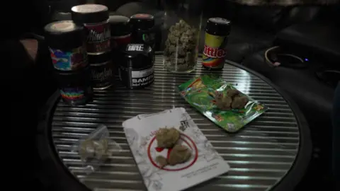 Tables showing plastic packets and jars bearing the names of "Cali" cannabis strains with skunk-style weed that was in them.